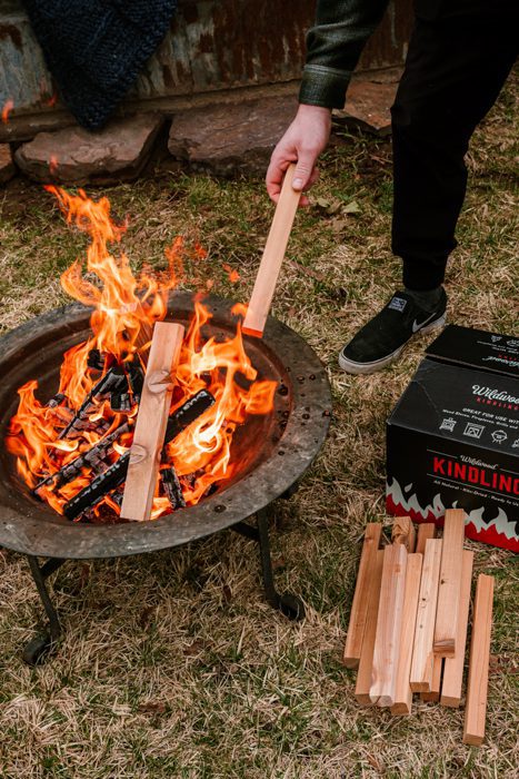 Person adding cedar kindling to a fire pit beside Wildwood Grilling kindling fire starters.
